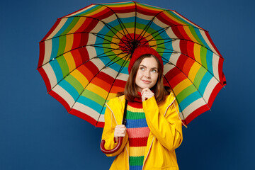 Young minded woman wear sweater red hat yellow waterproof raincoat outerwear hold opened colorful umbrella look aside isolated on plain dark blue background Outdoors wet fall weather season concept. - obrazy, fototapety, plakaty