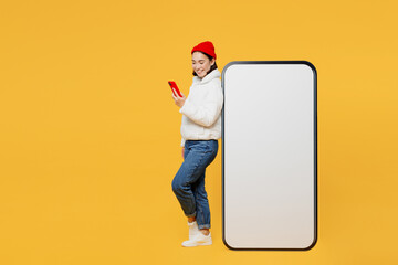 Full body side view fun young woman of Asian ethnicity in white padded windbreaker jacket red hat big huge blank screen mobile cell phone with area use smartphone isolated on plain yellow background