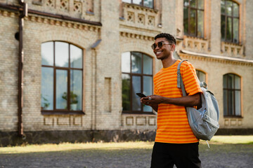 Young black man wearing sunglasses walking outdoors with cellphone