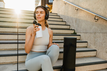 Young sportswoman drinking water while sitting on stairs after workout