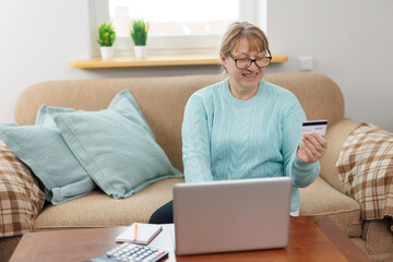 Clever senior satisfied woman sitting in the bright room on the sofa using the laptop and holding credit card.