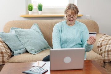 Clever senior satisfied woman sitting in the bright room on the sofa using the laptop and holding credit card.