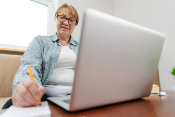 Happy mature businesswoman accountant in glasses and turquoise jumper sitting on the couch with laptop, taking notes, holding papers, contract studying or working remotely online from home