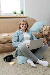Happy blond businesswoman with laptop looking at you while sitting on the floor of living-room and surfing in the net