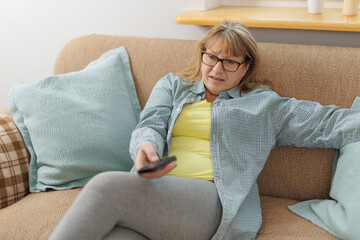 Obraz premium Mature blond woman watching television on sofa, in living room.