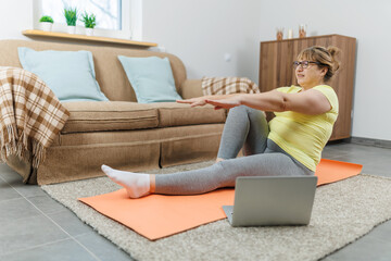 Mature woman exercising on the floor in front of laptop in the living room at home