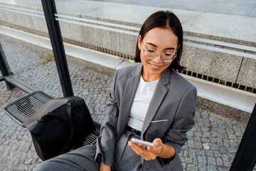Young woman smiling and using mobile phone while sitting at bus station