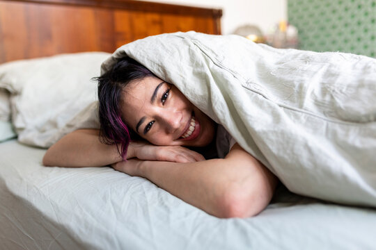 Happy young woman lying on bed with blanket at home