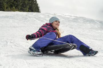 Happy little girl playing with a sled on a snowy hill. Girl enjoying a slide ride in the snow.