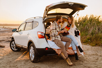 Happy young white couple smiling and sitting in car trunk outdoors