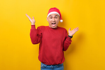 Fototapeta na wymiar Surprised young Asian man in Santa hat raising hands and looking at camera with excitement expression on yellow studio background. celebration Christmas holiday and New Year concept