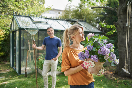Smiling woman smelling flower on potted plant with man in background at garden