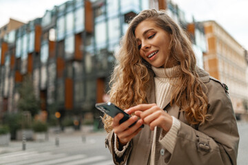 Smiling curly woman wearing warm coat walking down the street and using her phone