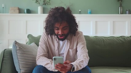 Young overjoyed Indian man with mobile phone makes victorious wave of hands and shouts with joy after reading news about winning of favorite team in baseball competitions sits on couch in apartment