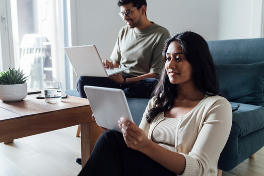 Young woman studying through tablet PC with man in background at home