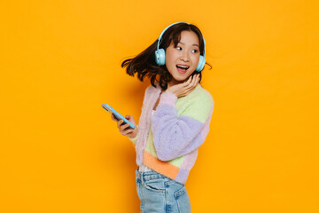 Asian woman in headphones dancing while listening music isolated over yellow background