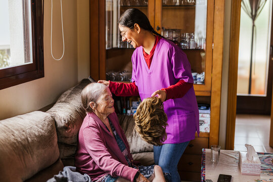 Happy young caregiver helping senior woman to put on wig at home