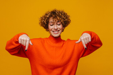 Cheerful middle-aged woman pointing fingers down isolated over yellow background