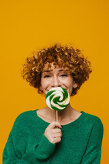 Middle-aged woman covering her mouth with lollipop over yellow background