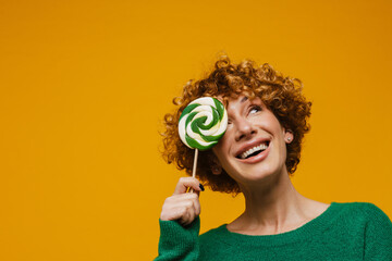 Middle-aged smiling woman covering her eye with lollipop isolated over yellow background
