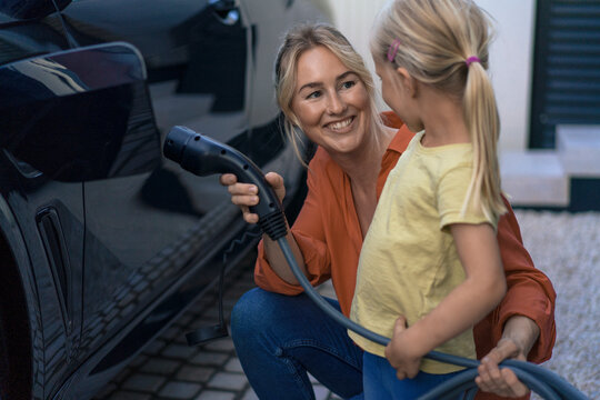 Smiling woman holding electric vehicle charger plug looking at daughter in front yard