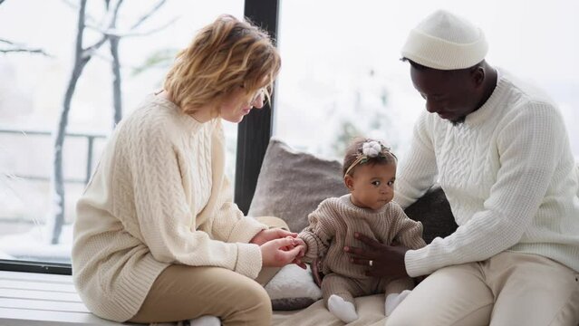 Happy interracial family sits on windowsill and plays with their baby daughter.