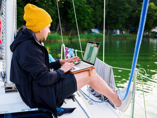 person wear jacket and knitted hat working with laptop on a sailing yacht during sailing by lake, remote or frilance work concept