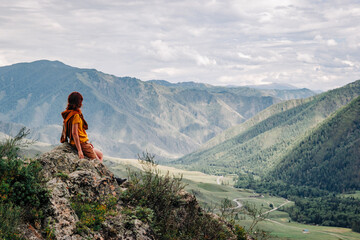 Side view of a red-haired girl sitting on a rock against the backdrop of a beautiful mountain valley