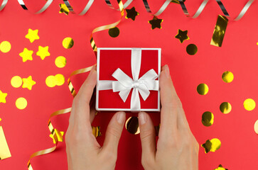 Red gift box with a white bow in female hands on a red background with serpentine and confetti
