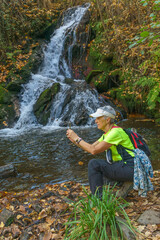 Naklejka na ściany i meble A woman takes a photo with her smartphone next to a flowing river in autumn, dressed in green sportswear. Hiking route in Aldeire, Granada, Spain.