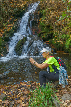 A woman takes a photo with her smartphone next to a flowing river in autumn, dressed in green sportswear. Hiking route in Aldeire, Granada, Spain.