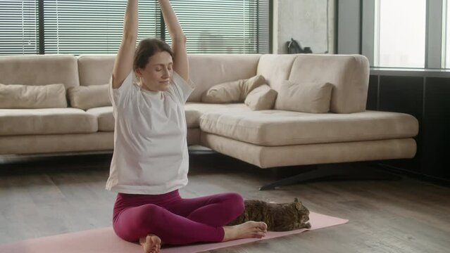 Middle age woman in sportswear practicing yoga at home with a cat at home. Female sit on mat in lotus position on red fitness mat in living room, healthcare and exercise mental therapy at home concept
