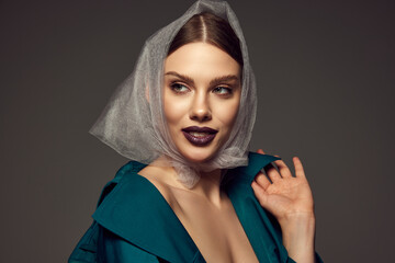 Portrait of young stylish woman with trendy dark lips make-up posing in fashionable coat and transparent headscarf over dark grey background. Stylish season