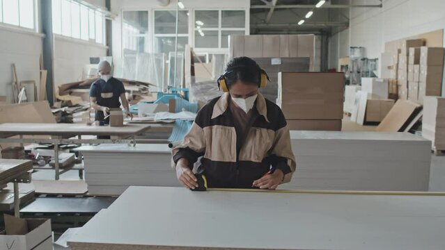 Female worker in protective ear muffs and mask measuring wooden board with tape and putting mark while working at furniture factory