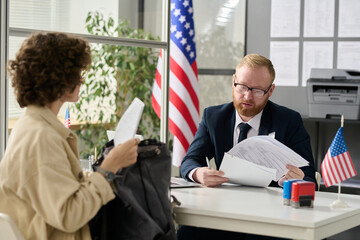 Portrait of male worker revising documents of young woman applying for visa in US immigration office - obrazy, fototapety, plakaty