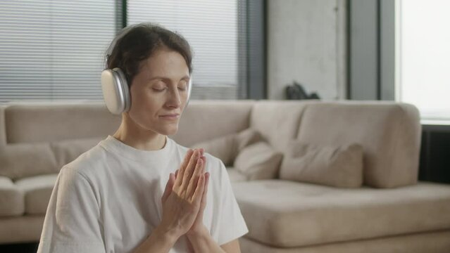 Middle age woman in sportswear practicing yoga at home. Girl sit on mat in lotus position on red fitness mat in living room with headphones with calm music. Female relaxing music for stress relief do
