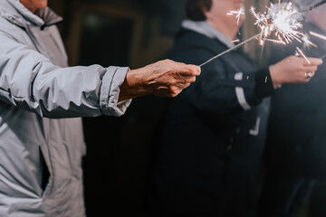 An old man holds a sparkling lights in his hand. Happy people with sparklers in their hands during celebration. Sparkler in hands on a wedding - guests holding lights in hand. 2023