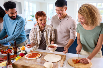 Group Of Friends At Home In Kitchen Adding Sauce To Homemade Pizzas For Party Together