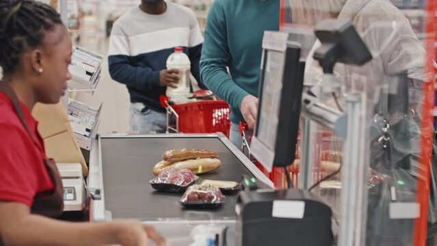 Queue of customers checking out at supermarket with female cashier sitting at cash counter and scanning their products