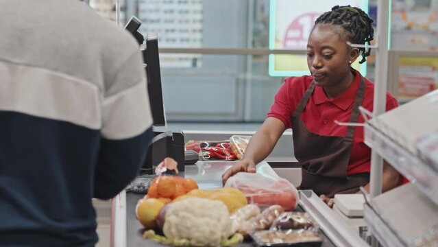 Young African American female cashier serving customer putting out his goods on conveyor belt at checkout in supermarket