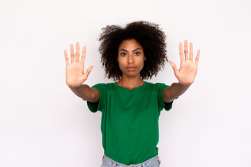 Portrait of serious young woman making stop gesture over white background. African American lady wearing green T-shirt and jeans showing restriction sign. Forbiddance concept