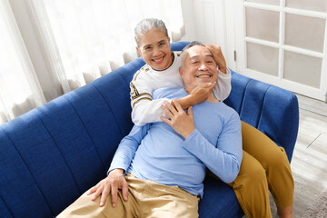 Portrait of happy Asian senior couple living together, hug, touching and embracing with a smile on the sofa in the living room. Retirement living together at home.