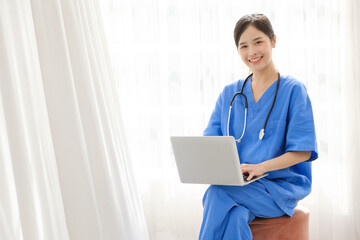 Portrait of a young Asian happy nurse wearing medical scrubs looking at the camera with a stethoscope and using a laptop while sitting with a white curtain in the background. Image with copy space.