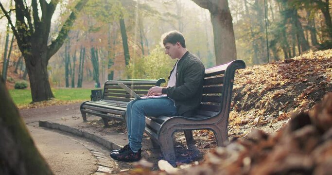 Caucasian young man sitting on bench in park, opening laptop and start working. Outdoor. Male freelancer typing on computer in nature in fall. Resting and chatting online. Remote work concept.