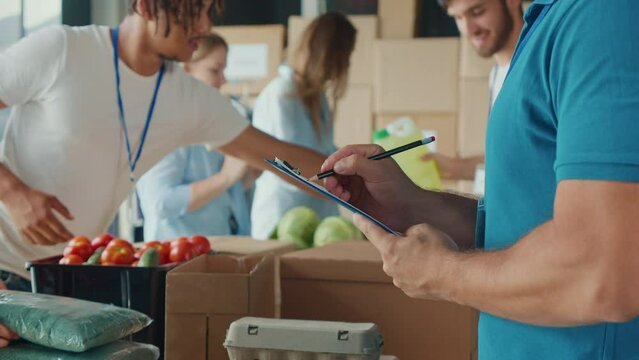 Close-up.Male Volunteer Writing on Clipboard While Working in Warehouse. Man Doing Inventory of Donated Goods in Volunteer Center While Group of Volunteers Sorting Humanitarian Aid on Background