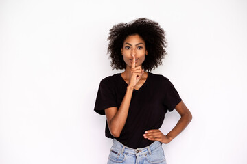 Fototapeta na wymiar Portrait of flirty young woman standing with finger in lips over white background. African American lady wearing black T-shirt and jeans making silence gesture. Secrecy concept