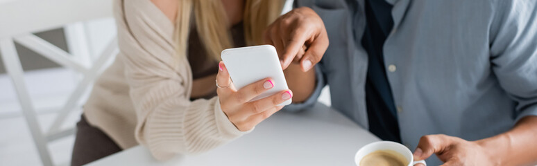 partial view of blurred african american man with coffee cup pointing at mobile phone near girlfriend in kitchen, banner