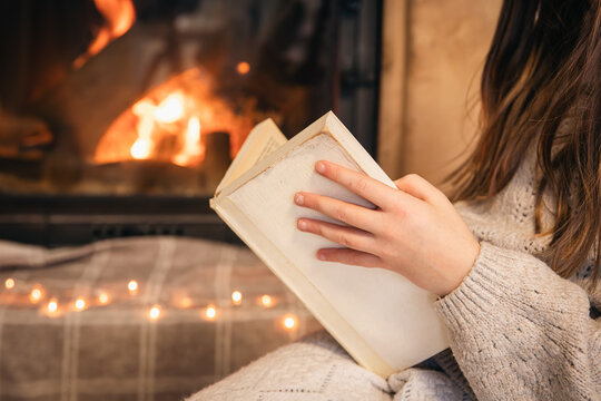 Little girl reading a book sitting by the fireplace at home, close-up.