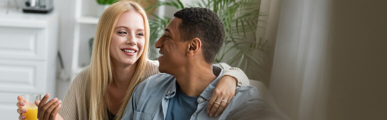smiling blonde woman holding orange juice and hugging african american boyfriend in kitchen, banner