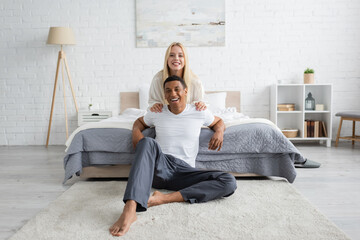 cheerful african american man in sleepwear sitting on floor in bedroom near blonde woman hugging...